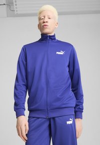 Lila, zipzáras sportdzseki sima anyagból, magas gallérral és fehér PUMA logóval a bal mellen. Passzoló nadrág látható.