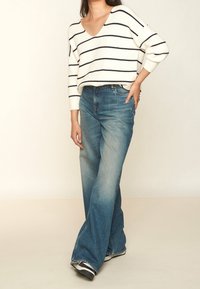 Pull à rayures blanches et bleu marine avec col en V, associé à un jean évasé bleu clair. La tenue met en avant un style décontract é, une coupe ample et des textures douces.