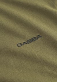 Olivovo zelená látka s hladkým povrchom, na ktorej sa v pravom dolnom rohu nachádza čierne vyšité logo "GABBA".