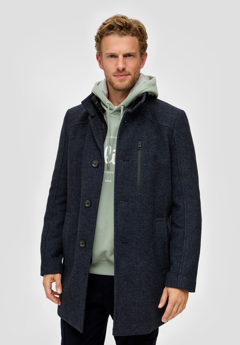 Donker marineblauwe wolblend jas met een hoge kraag, knoopsluiting, zijzakken en een ritsborstzak, gedragen over een lichtgroene hoodie.