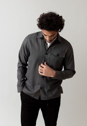 Camisa gris de botones con una tela texturizada, que incluye un bolsillo en el pecho y botones negros, combinada con pantalones negros.