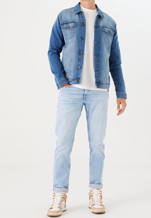 Mand iført en blå denimjakke, hvid t-shirt, lyseblå jeans og beige høje sneakers, stående foran en hvid baggrund.