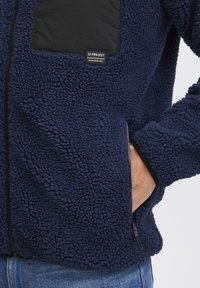 Navy fleece jacket with a textured surface, featuring a black chest pocket and a zippered side pocket. Includes a brand label on the pocket.