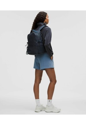 Granatowy plecak z wieloma przegrodami, prostokątny kształt, regulowane paski i teksturowane wykończenie. Noszony z szarym topem i niebieskimi szortami.