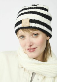 Zwart-wit gestreepte gebreide beanie met een textuurafwerking en een tan vierkante patch. Zachte crèmekleurige sjaal om de nek gedrapeerd.