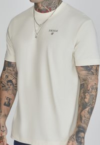 Kerman lyhythihainen t-paita, joka on valmistettu pehmeästä puuvillasta. Paitassa on pieni musta logo rinnassa ja rento istuvuus. Tatuoitu käsivarsi näkyvissä.