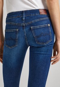 Jeans de mezclilla azul oscuro con un diseño ajustado, que cuentan con dos bolsillos traseros y un sutil detalle de costura. La tela texturizada presenta un acabado suave.