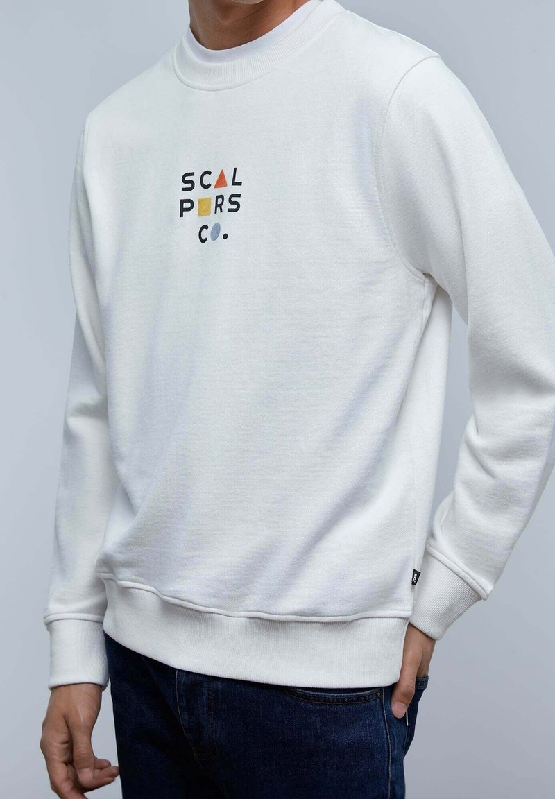 Homme portant un sweat-shirt blanc avec un texte abstrait coloré "SCAL PORS CO." et un jean bleu foncé, debout devant un fond clair.