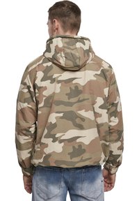 Veste de camouflage dans des tons beige et vert, dotée d'une capuche, de poignets élastiques et d'une coupe décontractée. Le matériau semble lisse et léger.