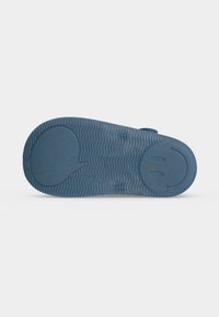 Semelle de chaussure bleue avec un motif ondulé, une grande zone circulaire au talon avec un visage souriant, et une forme de bulle au niveau de la pointe.