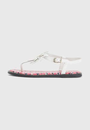 Hvid flad sandal med blomsterkvastdetalje, justerbar ankelrem og lyserød mønstret sål på gråt baggrund.
