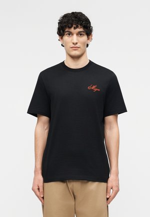 Νεαρός με σγουρά σκούρα μαλλιά φοράει μαύρο T-shirt με μικρό κόκκινο λογότυπο σε στυλ γραφικού στο στήθος και μπεζ παντελόνι, στέκεται μπροστά σε απλό φόντο.