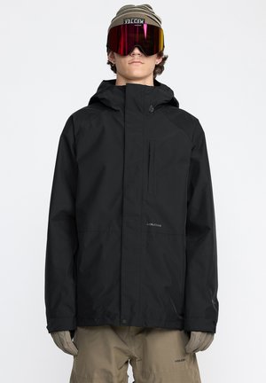 Veste imperméable noire avec une capuche, zipper avant et poche sur la poitrine. Portée avec un pantalon beige et des lunettes de protection réfléchissantes surdimensionnées.