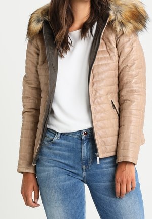 Femme portant une veste matelassée beige avec une capuche bordée de fourrure, une chemise blanche et un jean bleu, posant devant un fond uni.