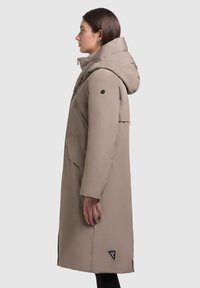 Dlhý béžový nepremokavý kabát s kapucňou, predným zipsom, gombíkovými akcentmi a trojuholníkovou nášivkou blízko lemu. Má štruktúrovaný siluet.