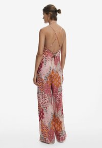 Mono multicolor con espalda cruzada, que presenta patrones florales y geométricos en tonos de rojo, rosa y naranja; piernas anchas y fluidas.