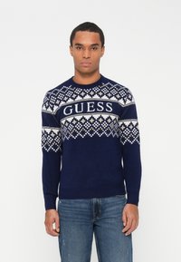 Maglione lavorato a maglia blu navy con un motivo geometrico in crema e grigio chiaro, con il logo "GUESS" sul petto. Maniche lunghe e collo rotondo.