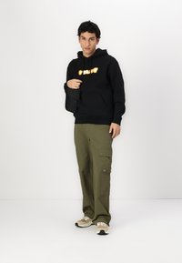 Svart hettegenser med oransje tekst, olivengrønne cargobukser og beige joggesko. Hettegenseren har en frontlomme og justerbare snorer.