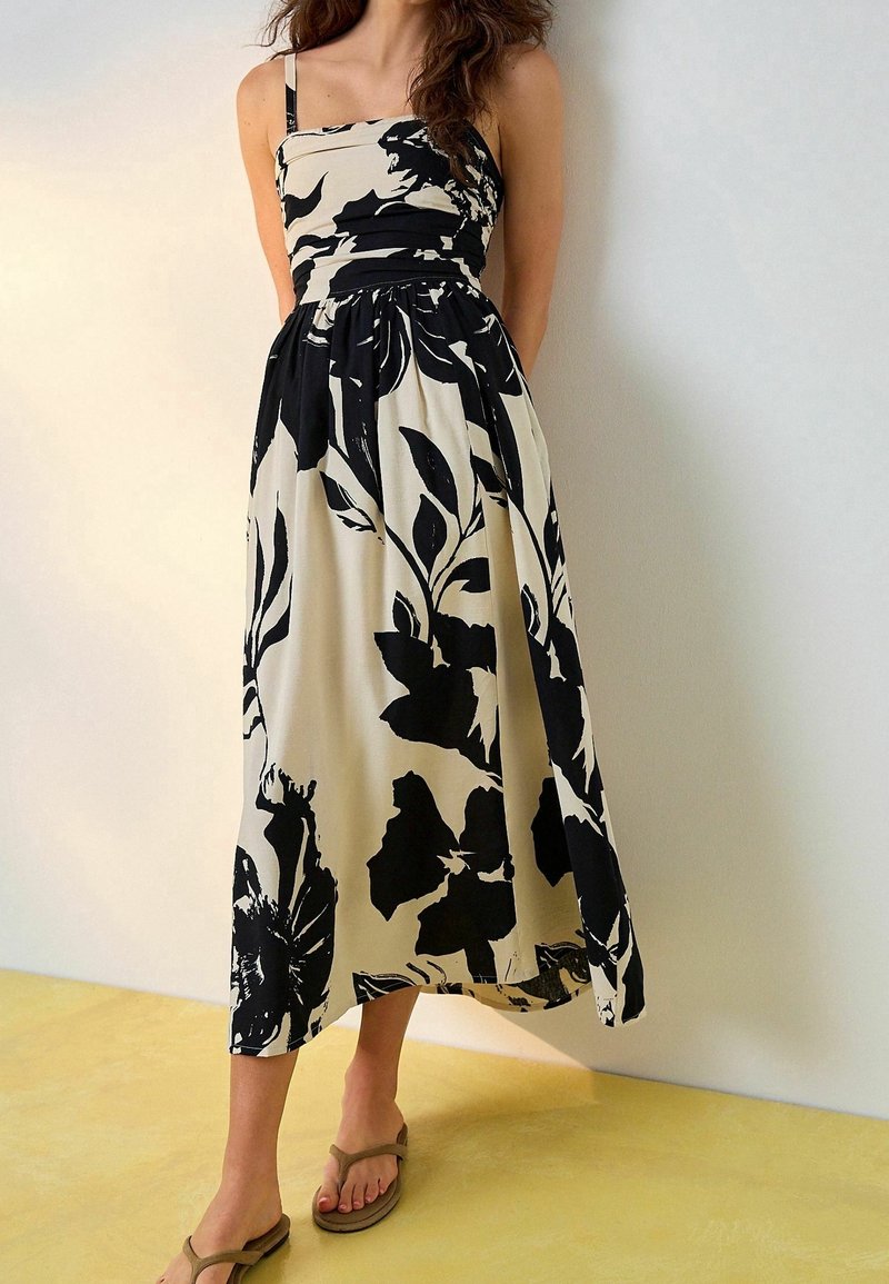 Femme portant une robe midi fleurie noire et blanche sans manches avec des fines bretelles et des sandales beiges, debout contre un mur uni.