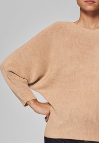 Beige oversized gebreide trui met een brede halslijn en lange, losse mouwen. Gestructureerde stof met een geribbelde patroon.