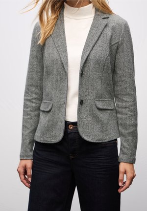 Vrouw draagt een grijs visgraatblazer over een witte coltrui en donkere spijkerbroek, staand tegen een effen lichte achtergrond.