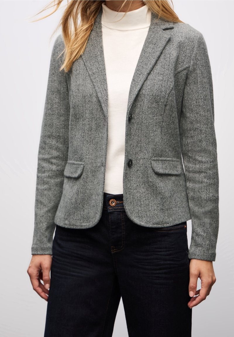 Femme portant un blazer gris en chevrons par-dessus un col roulé blanc et un jean foncé, debout devant un fond clair uni.