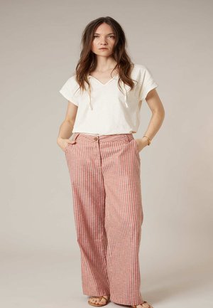 Femme portant un haut blanc à manches courtes avec col en V et cordons, pantalon large rayé rose et blanc, les mains dans les poches, debout devant un fond uni.