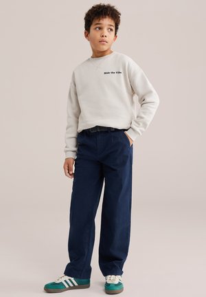 Zēns ar cirtainiem matiem, valkā bēša krāsas džemperi, tumši zilas bikses un zaļus sporta apavus, stāv ar vienu roku kabatā un skatās sāniski.