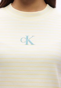 Λευκή και ανοικτοκίτρινη ριγωτή μπλούζα με ριμπ ύφανση, διακοσμημένη με τυρκουάζ λογότυπο "CK" στο στήθος. Στρογγυλός λαιμός.