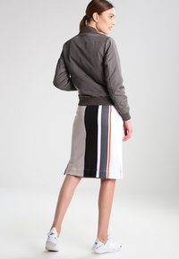Graue Bomberjacke mit gerippten Bündchen, kombiniert mit einem knielangen Rock mit vertikalen Streifen in Grau, Schwarz und Weiß, ergänzt durch rote Akzente.
