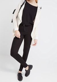 Cardigan léger en crème avec un devant ouvert, associé à un haut noir et un jean noir ajusté. Des chaussures noires à lacets complètent la tenue.
