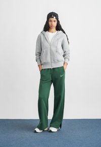 Jeune personne portant une casquette noire à l'envers, un sweat à capuche gris zippé, un pantalon de jogging vert Nike et des baskets blanches, se tenant contre un mur blanc uni.