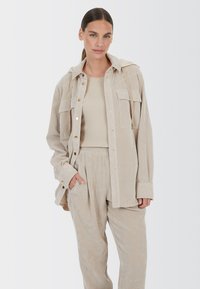 Beige corduroy shirt jacket with two front pockets, gold buttons, and a hood, paired with matching ribbed tank top and loose-fit pants.