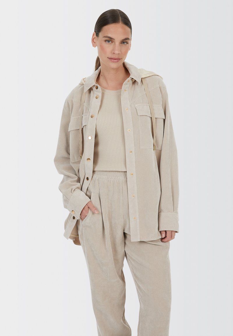 Beige corduroy shirt jacket with two front pockets, gold buttons, and a hood, paired with matching ribbed tank top and loose-fit pants.