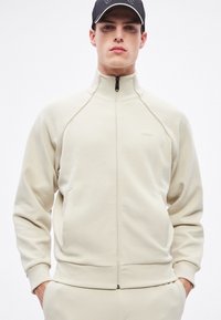 Kremowa bluza na zamek z raglanowymi rękawami, bocznymi kieszeniami, elastycznymi mankietami i gładką fakturą; logo wytłoczone na piersi.