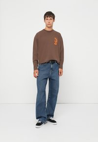 Ung mand iført en brun langærmet skjorte med orange logo, løse blå jeans, sort bælte og sorte og hvide sneakers, stående mod en hvid væg.
