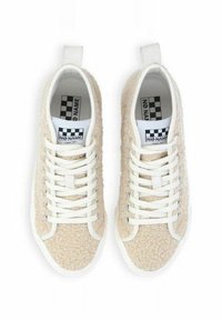 Zapatillas beige de textura esponjosa con cordones blancos, suelas de goma y una lengüeta. Presenta una etiqueta a cuadros en el interior con el nombre de la marca.