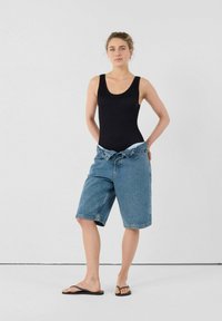 Zwart geribbeld body, gecombineerd met lichtblauwe denimshorts, tot boven de knie. Model staat op blote voeten met handen op de heupen tegen een neutrale achtergrond.