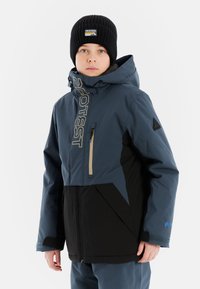 Blauwe en zwarte skijas met een hoge kraag, een ritsborstzak, logo op de voorkant en een geometrisch ontwerp op de schouders. Draagt een zwarte beanie.