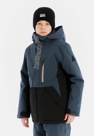 Blauwe en zwarte skijas met een hoge kraag, een ritsborstzak, logo op de voorkant en een geometrisch ontwerp op de schouders. Draagt een zwarte beanie.