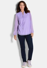 Blusa viola chiaro con maniche lunghe trasparenti e scollatura a goccia, abbinata a pantaloni blu scuro e sneakers viola chiaro.