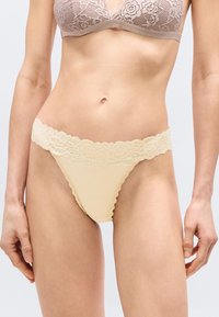 Γυναίκα που φορά σετ εσωρούχων σε μπεζ με δαντέλα, παρουσιάζοντας το λεπτομερές λουλουδένιο μοτίβο δαντέλας στο σουτιέν και τα ταιριαστά εσώρουχα με ψηλή κοπή.