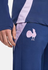 Haut de sport bleu avec des accents violet clair, comportant un logo de coq rose en bas à droite, mettant en valeur un tissu texturé.