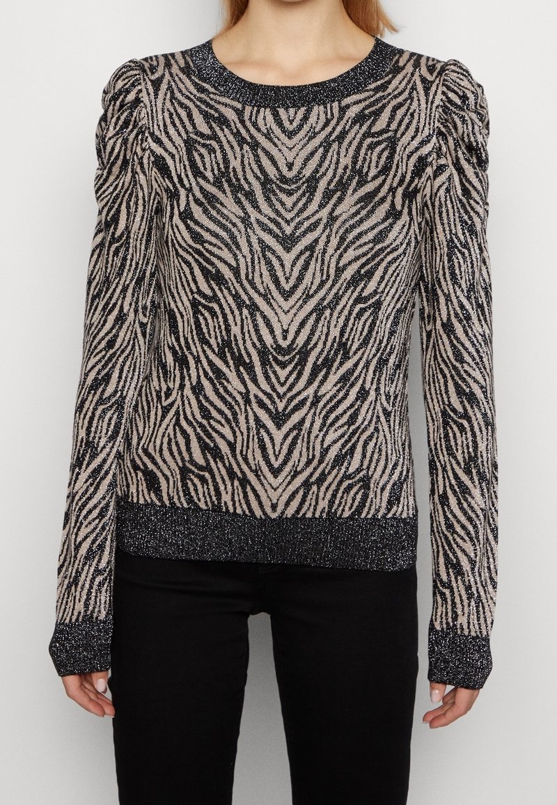 Femme portant un pull ajusté à manches longues avec des rayures zébrées noires et beiges ainsi que des poignets, un ourlet et un col noirs pailletés.