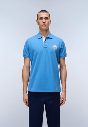 Homme debout portant un polo bleu clair avec un petit logo rond sur la poitrine et un pantalon bleu foncé, sur un fond uni.