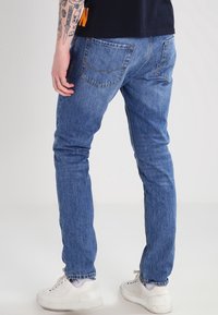 Person, der blaue Jeans und weiße Sneaker trägt, mit dem Rücken zur Kamera steht und Tätowierungen am linken Arm zeigt.