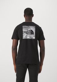 Muž oblečený v černém tričku s bílou abstraktní grafikou hory a slunce a logem "The North Face" na zadní straně, stojící zády.