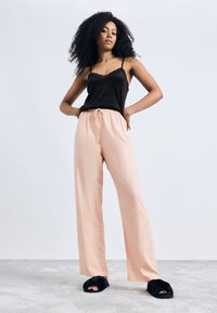 Zwarte top met kant, perzikkleurige broek met trekkoord en zwarte pluizige slides. Model staat met handen op de heupen tegen een lichte achtergrond.