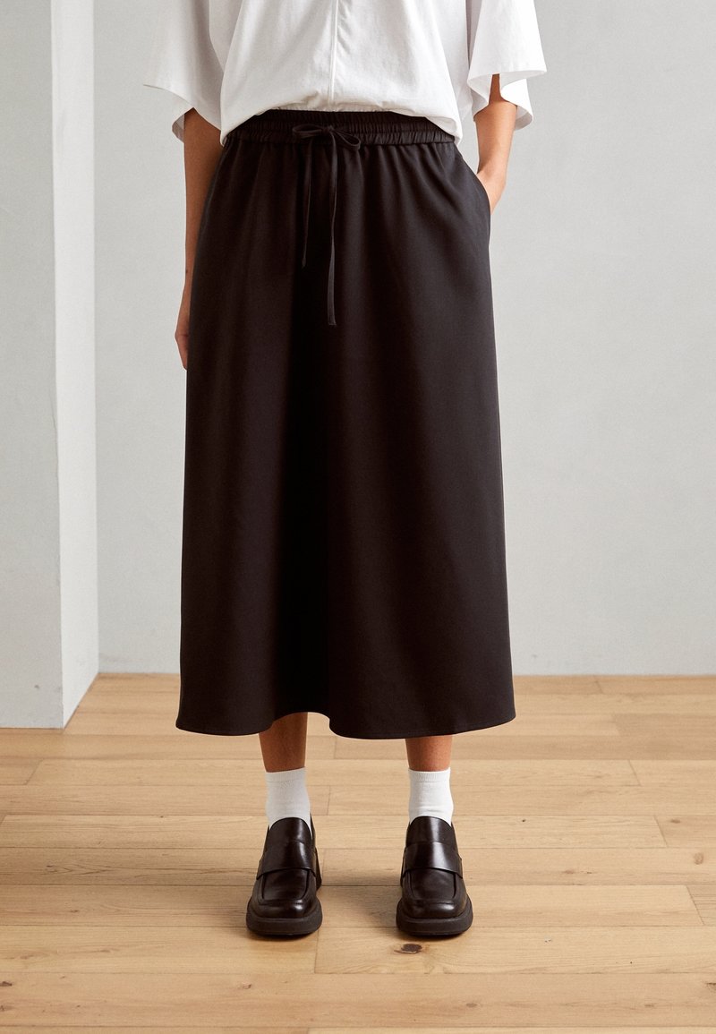 Personne portant un haut blanc, une jupe midi noire avec une taille à cordon, des chaussettes blanches et des mocassins noirs debout sur un sol en bois.