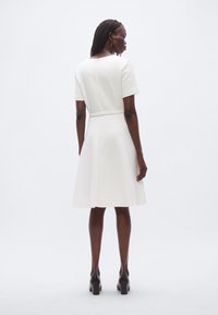 Robe blanche texturée avec manches courtes, ceinture à la taille et jupe évasée. Dotée d'une ouverture en forme de trou de serrure dans le dos. Associée à des chaussures à talons noires.